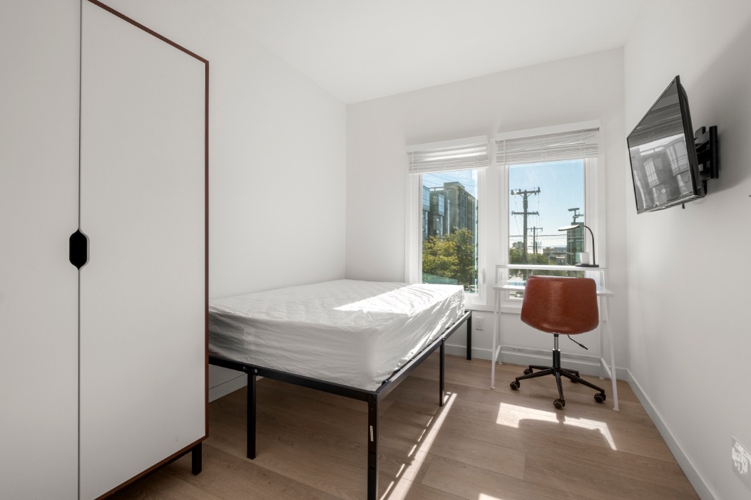 Bright and modern bedroom with a large window offering a city view, sleek white wardrobe, comfortable bed, and stylish work desk with chair.