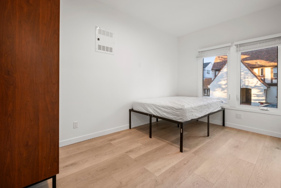 Bright minimalist bedroom with large window, offering a view of charming architecture and featuring light hardwood floors and a cozy bed.