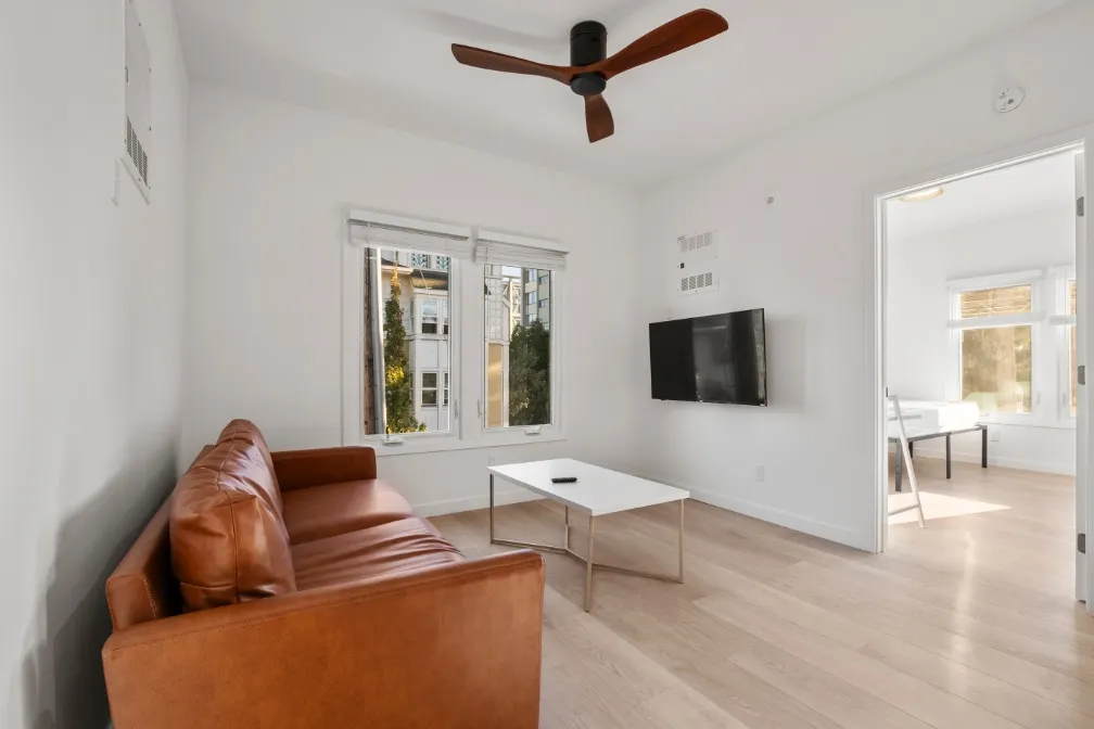 Bright and modern living room with a sleek leather sofa, mounted flat-screen TV, and large windows offering abundant natural light.