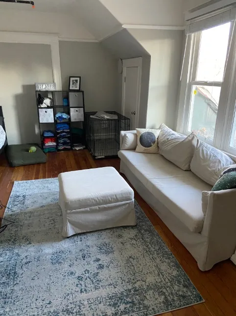 Charming living room with cozy white sofa, stylish ottoman, and natural light through large windows.