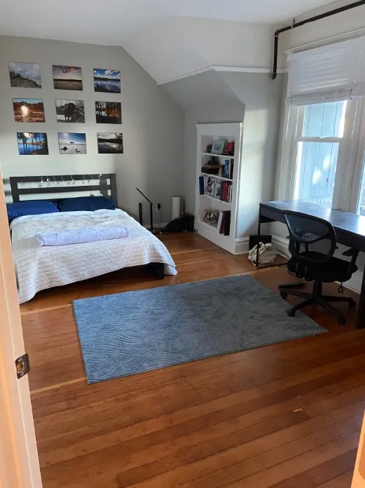Charming bedroom with wooden floors, stylish wall decor, cozy bedding, and a work-friendly desk near a large window.