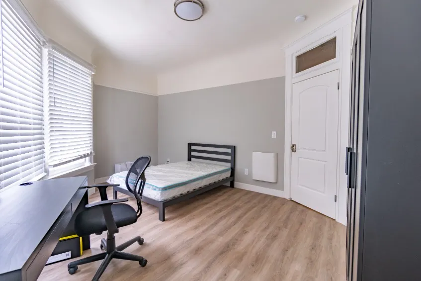 Bright, modern bedroom featuring a sleek desk, comfortable office chair, and a cozy bed, enhanced by natural light from large windows with blinds.