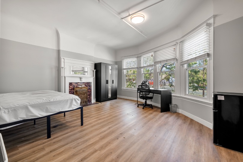 Bright and spacious bedroom with stylish hardwood floors, featuring a cozy bed, modern black wardrobe, and a functional desk area by large bay windows.