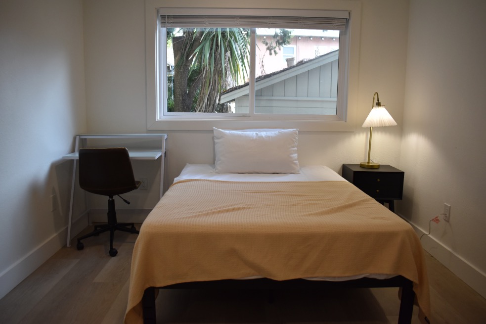 Bright and cozy bedroom with a comfortable bed, elegant lamp, and a window view of lush greenery.