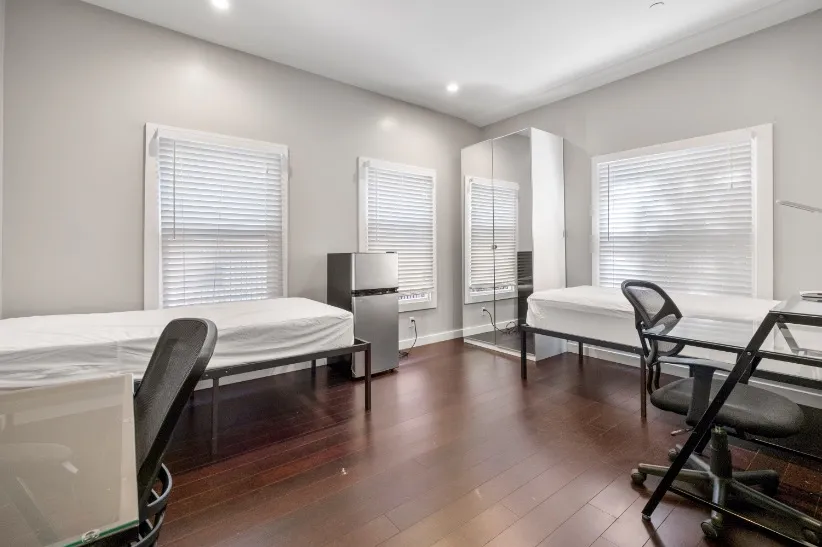 Modern, well-lit bedroom featuring two beds, sleek hardwood flooring, and large windows with blinds, creating a bright and inviting atmosphere.