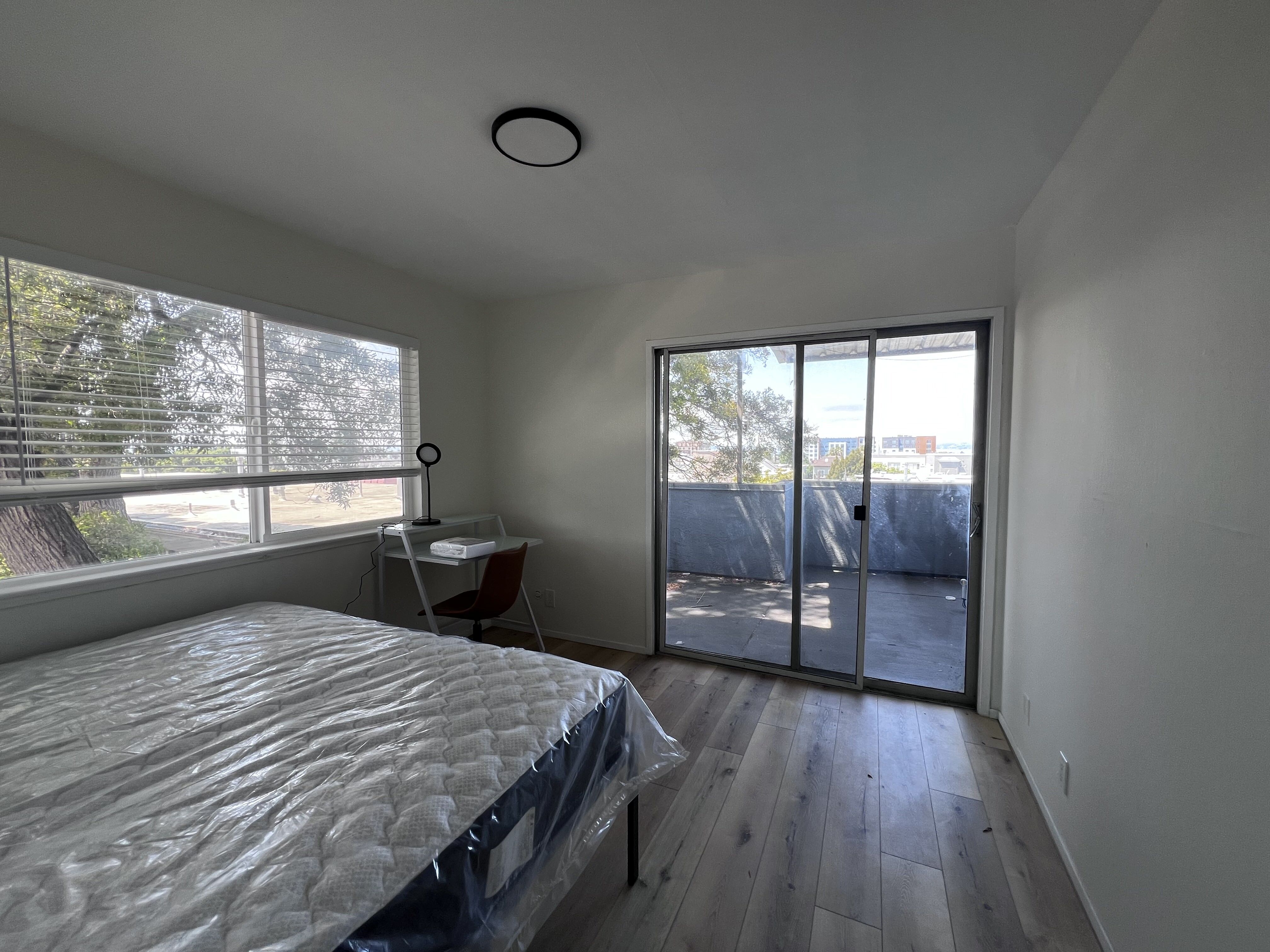 Bright and airy bedroom with large windows and sliding glass doors leading to a private balcony, featuring a cozy workspace and modern wood flooring.