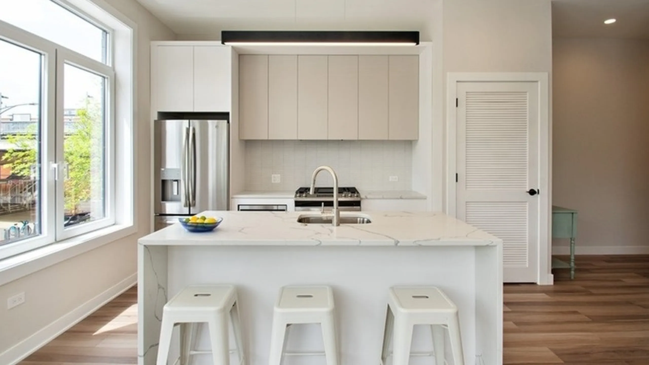 Contemporary kitchen with sleek white cabinets, marble countertop island, and stainless steel appliances illuminated by large windows.
