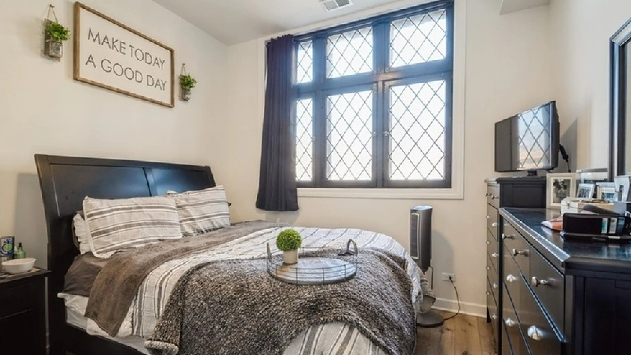 Bright, cozy bedroom featuring a plush bed with elegant linens, large window with charming lattice design, and modern dresser, creating a warm and inviting atmosphere.