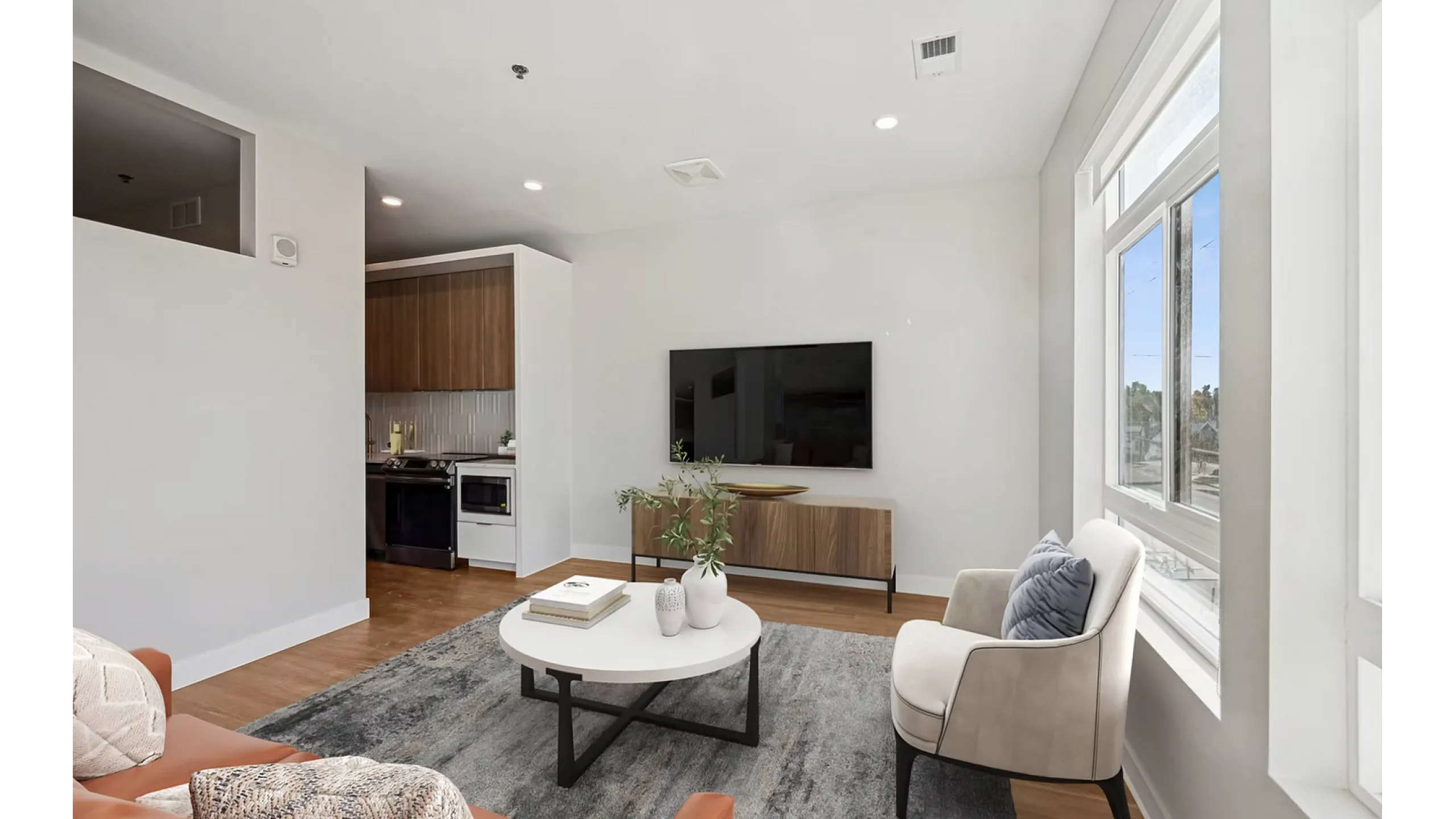 Bright and modern living room with sleek wooden flooring, featuring a cozy seating area, elegant decor, large flat-screen TV, and abundant natural light from expansive windows.