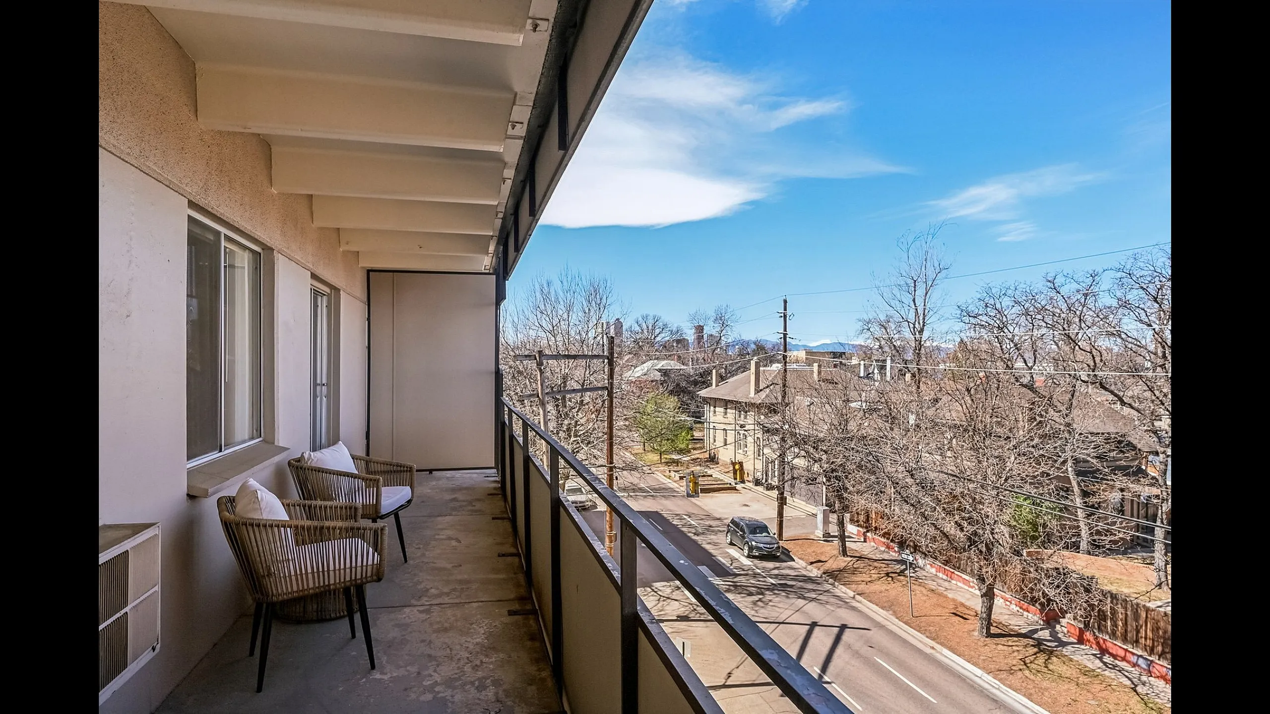 Expansive balcony view with cozy seating, overlooking a vibrant neighborhood street and clear blue skies.