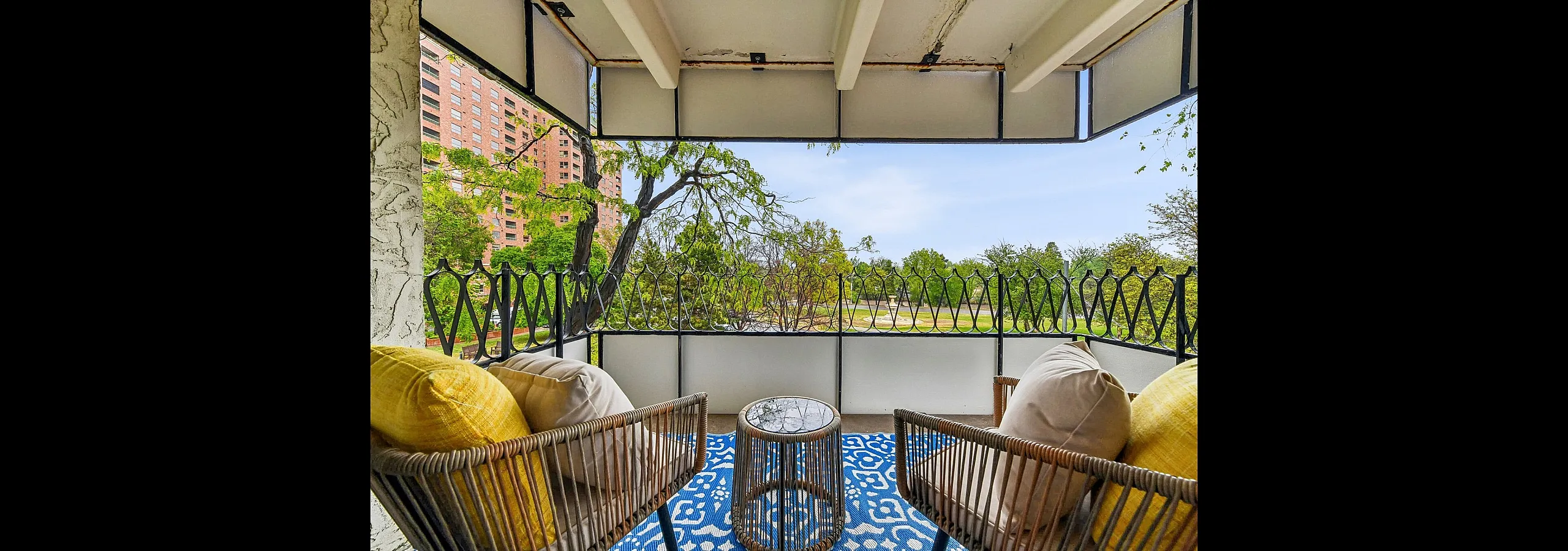 Charming balcony with stylish wicker seating, vibrant cushions, and a scenic view of lush greenery and a distant red-brick building.