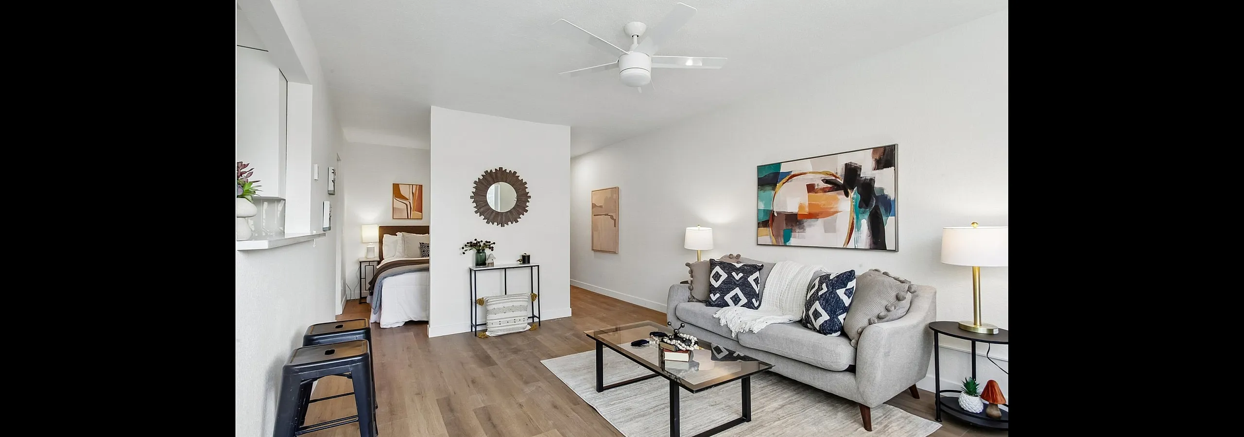 Elegant living room with sleek wood flooring, modern art on walls, and plush gray sofa with decorative pillows, adjacent to a cozy bedroom, creating a warm and inviting open space.