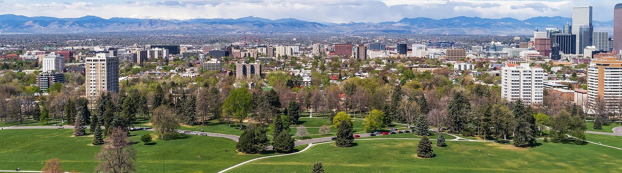 Stunning cityscape view showcasing lush green parks, tall buildings, and a backdrop of majestic mountains under a vibrant blue sky.