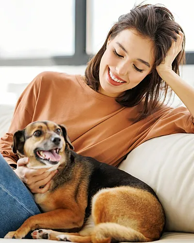 Bright, cozy living room with large windows, featuring a happy woman relaxing on a comfortable cream sofa with her dog.