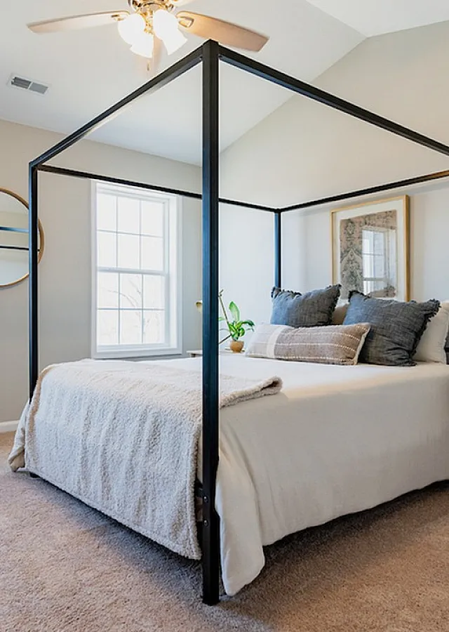 Bright and cozy bedroom with a stylish canopy bed, plush carpeting, and ample natural light from large windows, creating a serene and welcoming atmosphere.