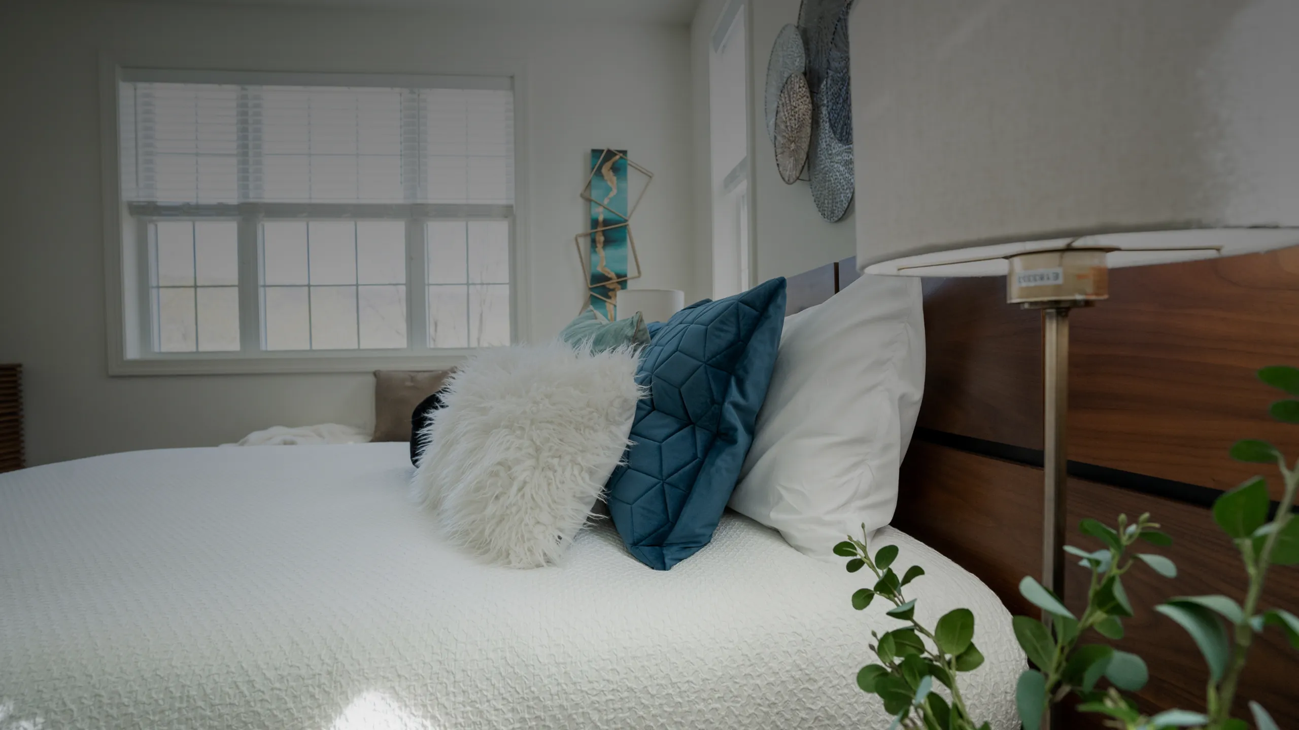 Sophisticated bedroom with a cozy ambiance, featuring a plush white bedspread, vibrant teal and fluffy white pillows, elegant wall art, and natural light streaming through large windows.