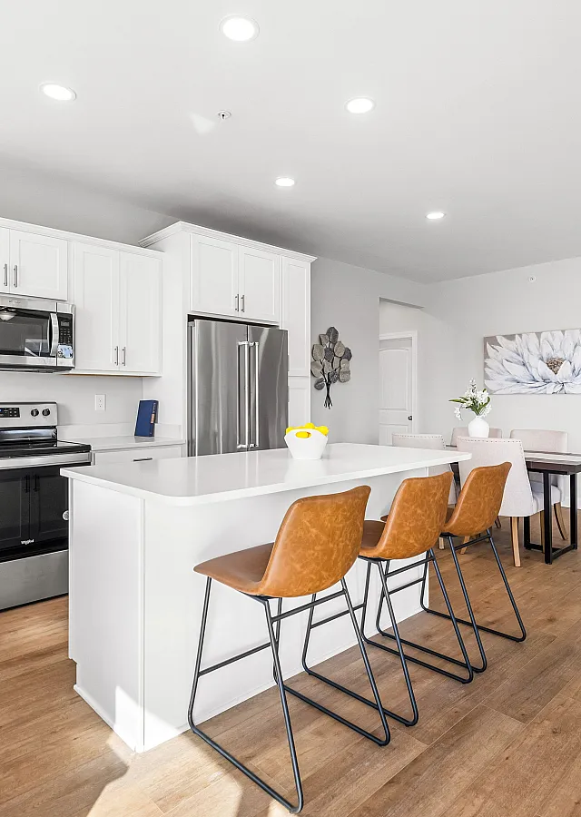 Bright and modern kitchen with sleek white cabinetry, stainless steel appliances, and a stylish breakfast bar featuring leather stools, leading to an elegant dining area.