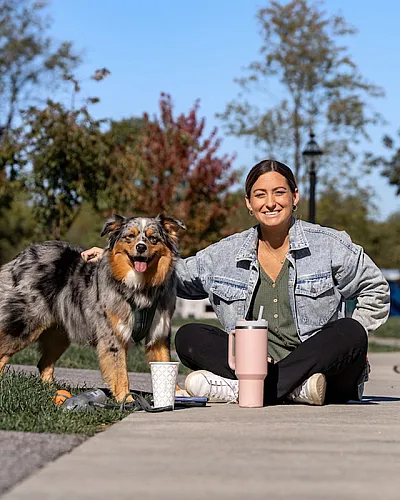 Woman enjoying a sunny day sitting on a pathway with her happy dog beside her, surrounded by lush greenery and clear blue skies.