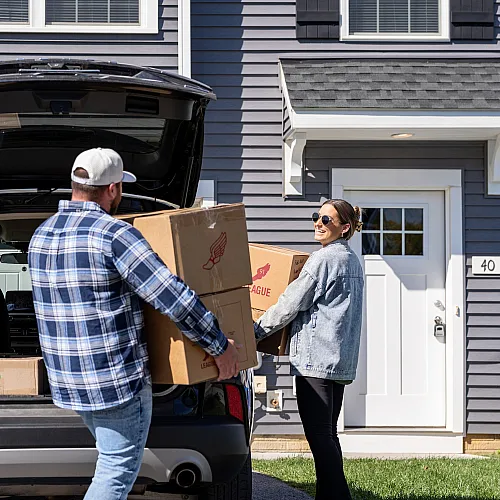 Cheerful couple moving into a modern townhouse, unloading boxes from a car on a sunny day.