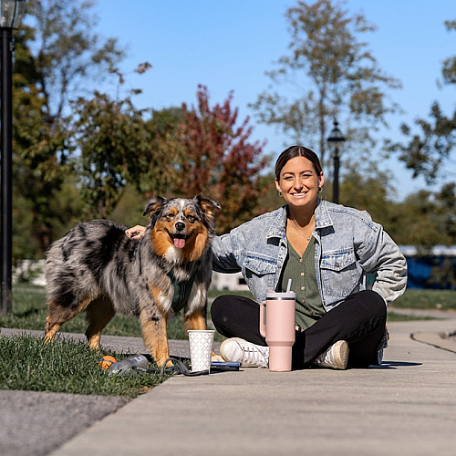 Woman enjoying a sunny day in the park with her playful dog, sitting on the grass with a refreshing beverage.