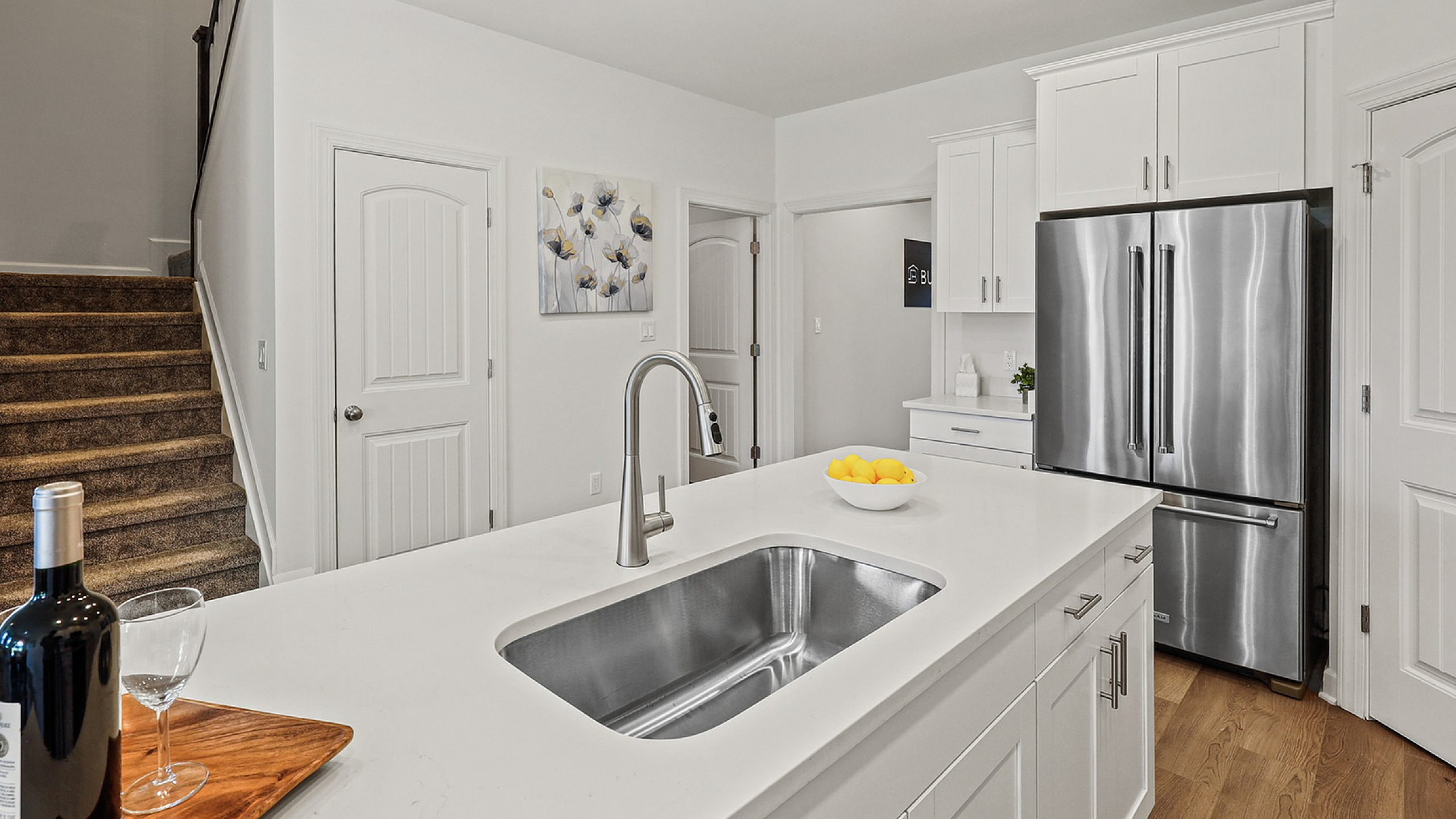 Modern kitchen with sleek white cabinetry, stainless steel appliances, and a stylish island featuring a wine bottle and fruit bowl, leading to a carpeted staircase.