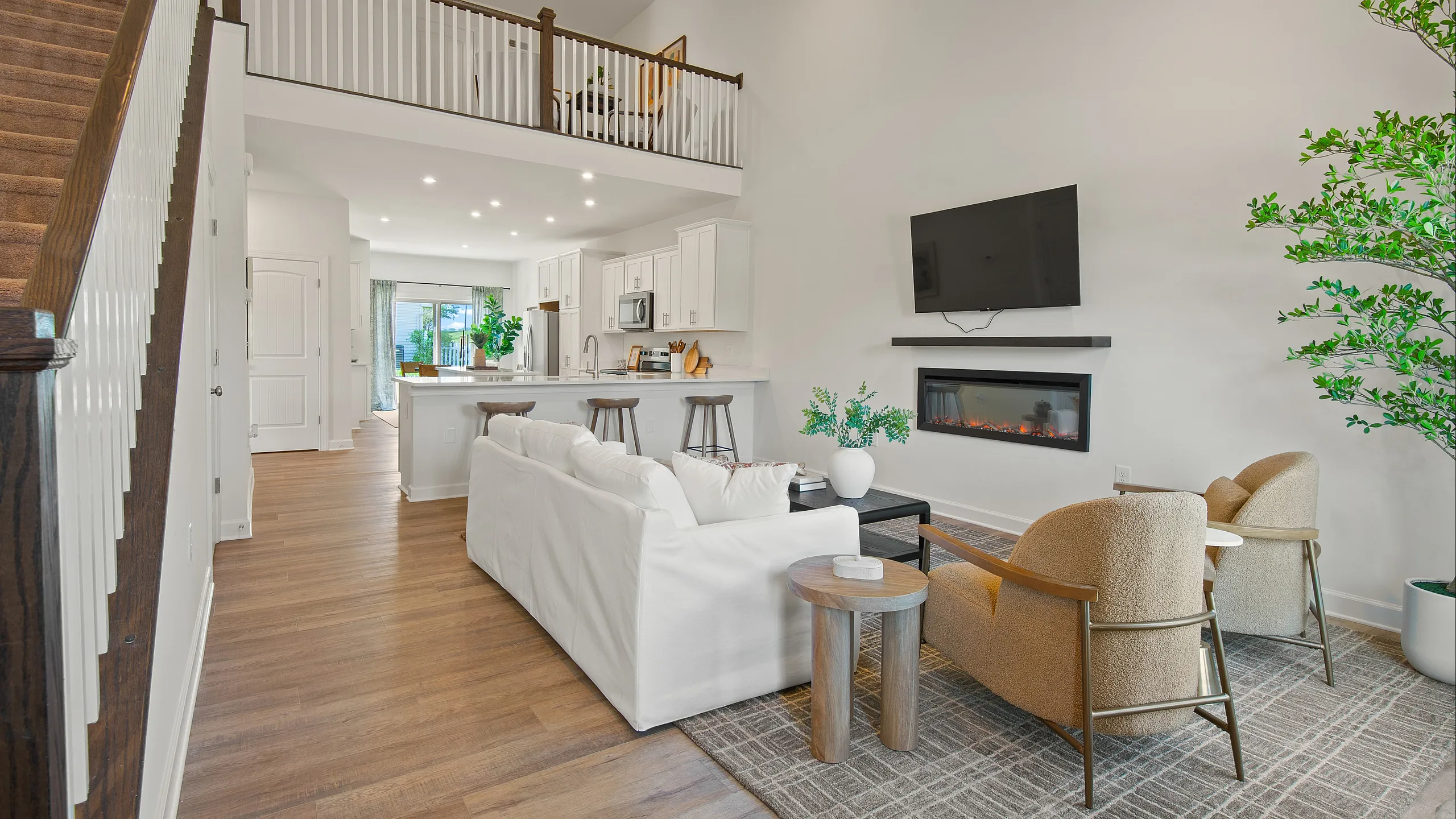 Bright and inviting open-concept living space featuring a modern kitchen with bar seating, stylish living room with a cozy fireplace, and an elegant staircase leading to an upper loft area.