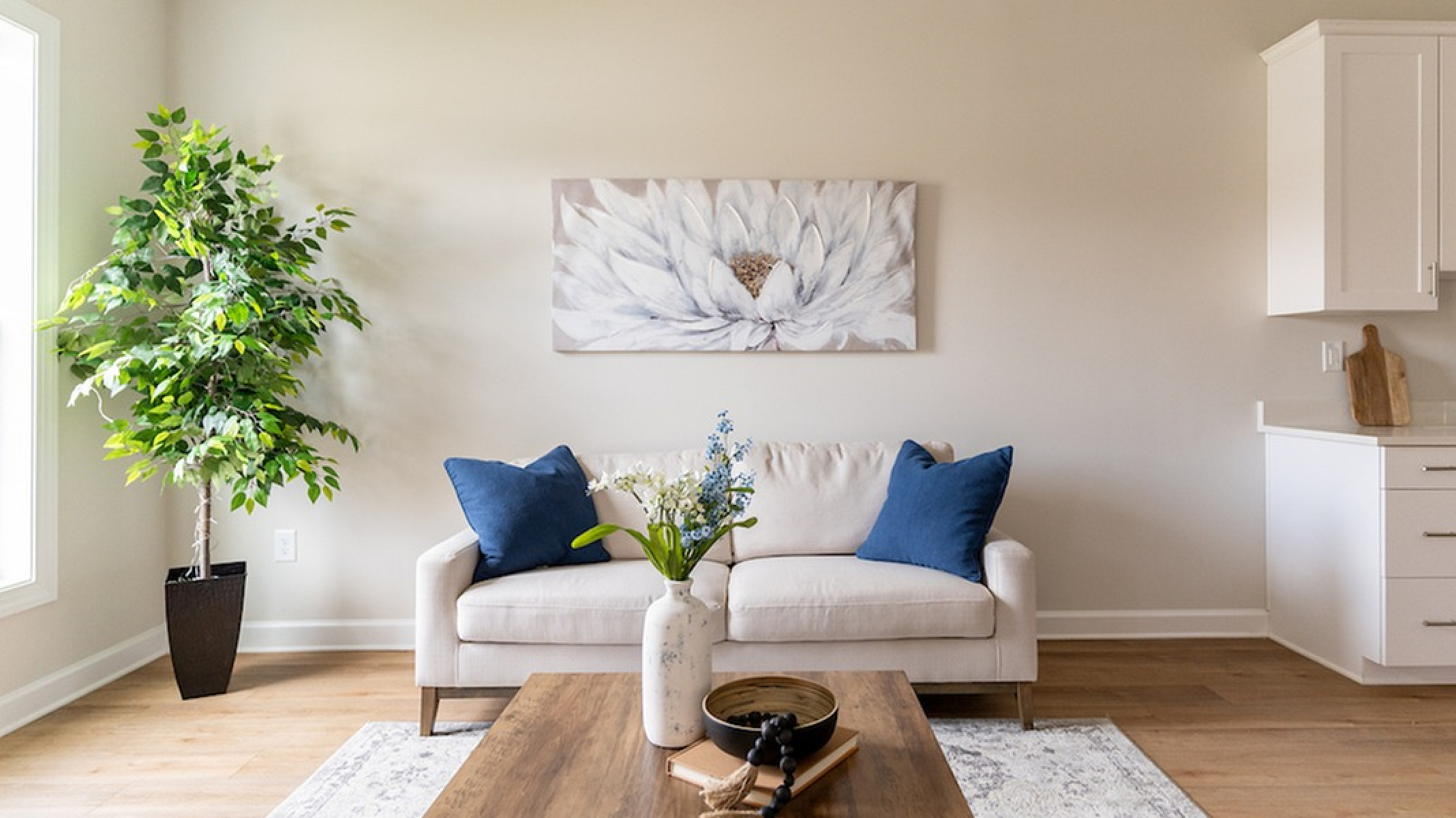 Bright and inviting living room with elegant decor, featuring a cozy cream sofa with blue accent pillows, a chic wooden coffee table, a lush indoor plant, and a modern kitchen area.