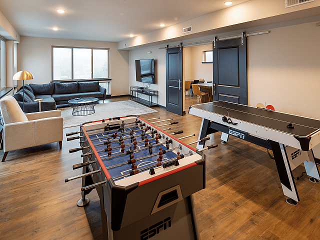 Spacious entertainment room with sleek foosball and air hockey tables, stylish seating area with a modern sofa, and large flat-screen TV in a beautifully lit space.