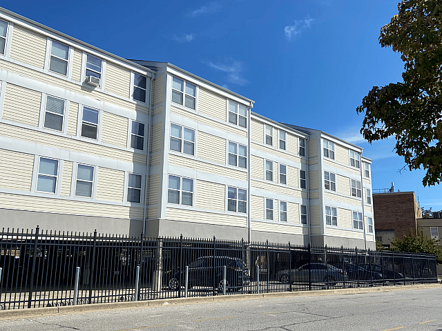 Charming multi-story apartment building with spacious windows, surrounded by a secure fence against a clear blue sky.