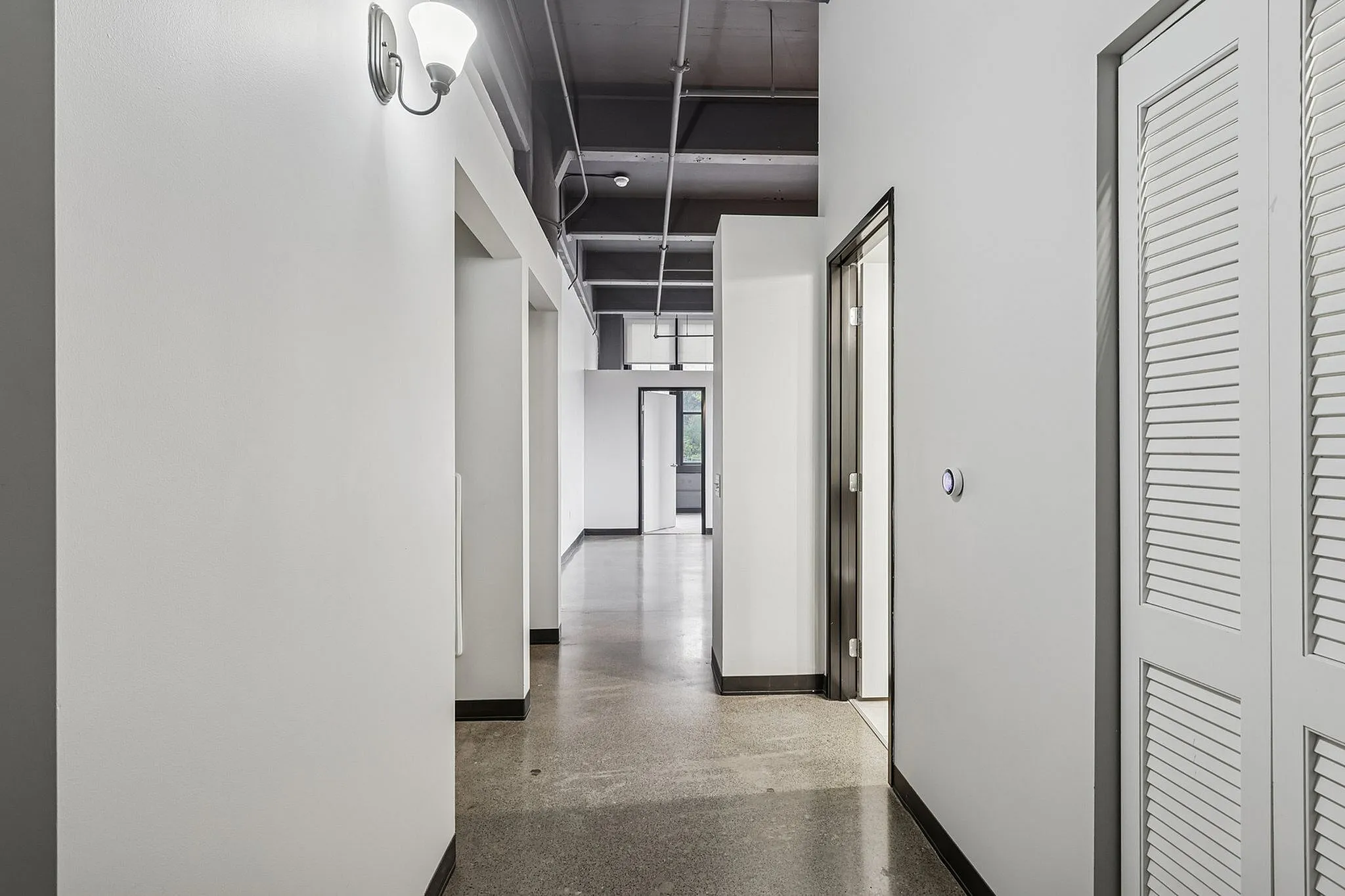 Sleek modern hallway with polished concrete floors, industrial exposed ceiling, and bright natural light leading to open-plan living spaces.