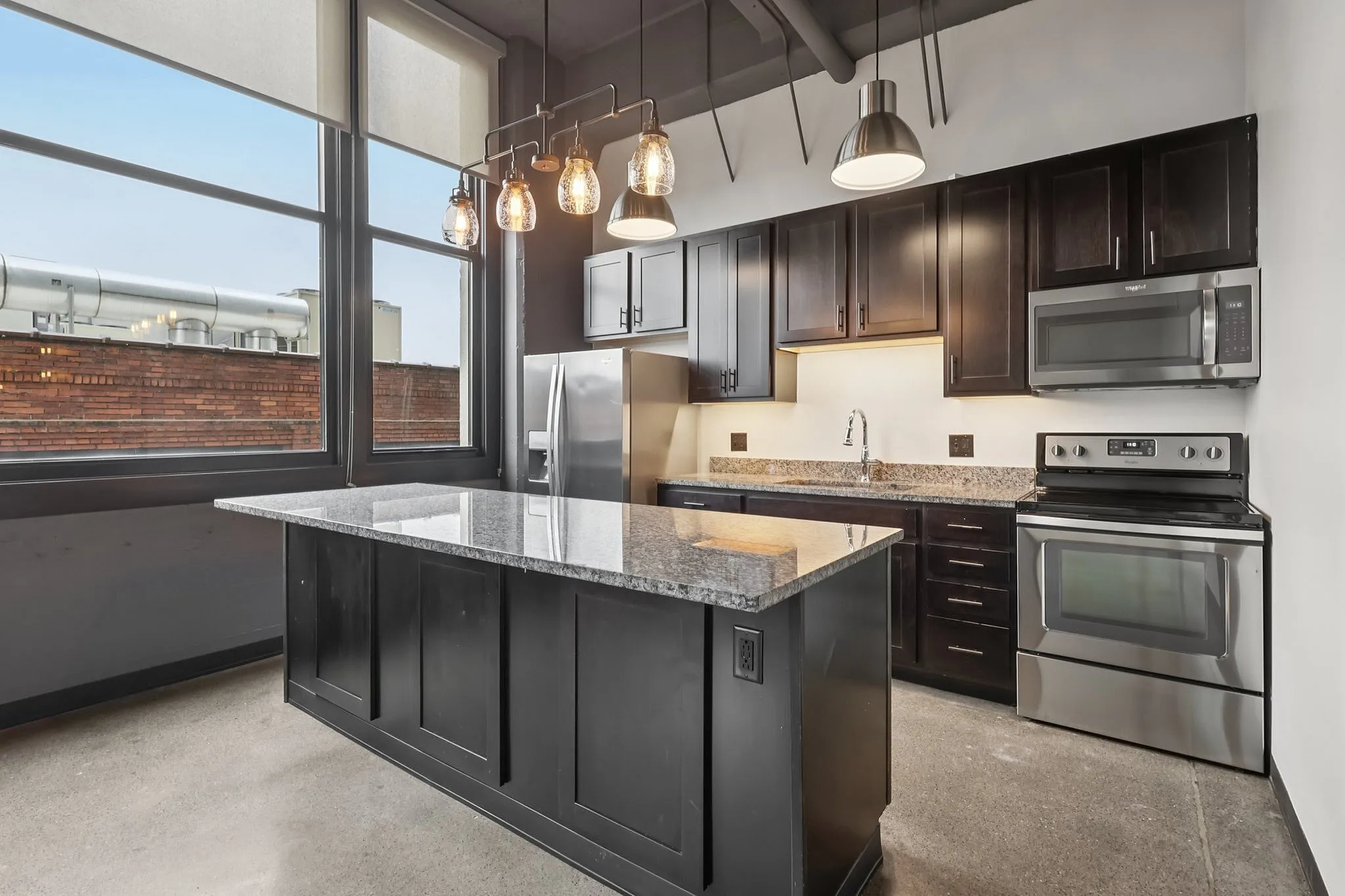 Modern industrial kitchen with sleek dark cabinets, stainless steel appliances, and stylish pendant lighting, offering a sophisticated cooking environment.