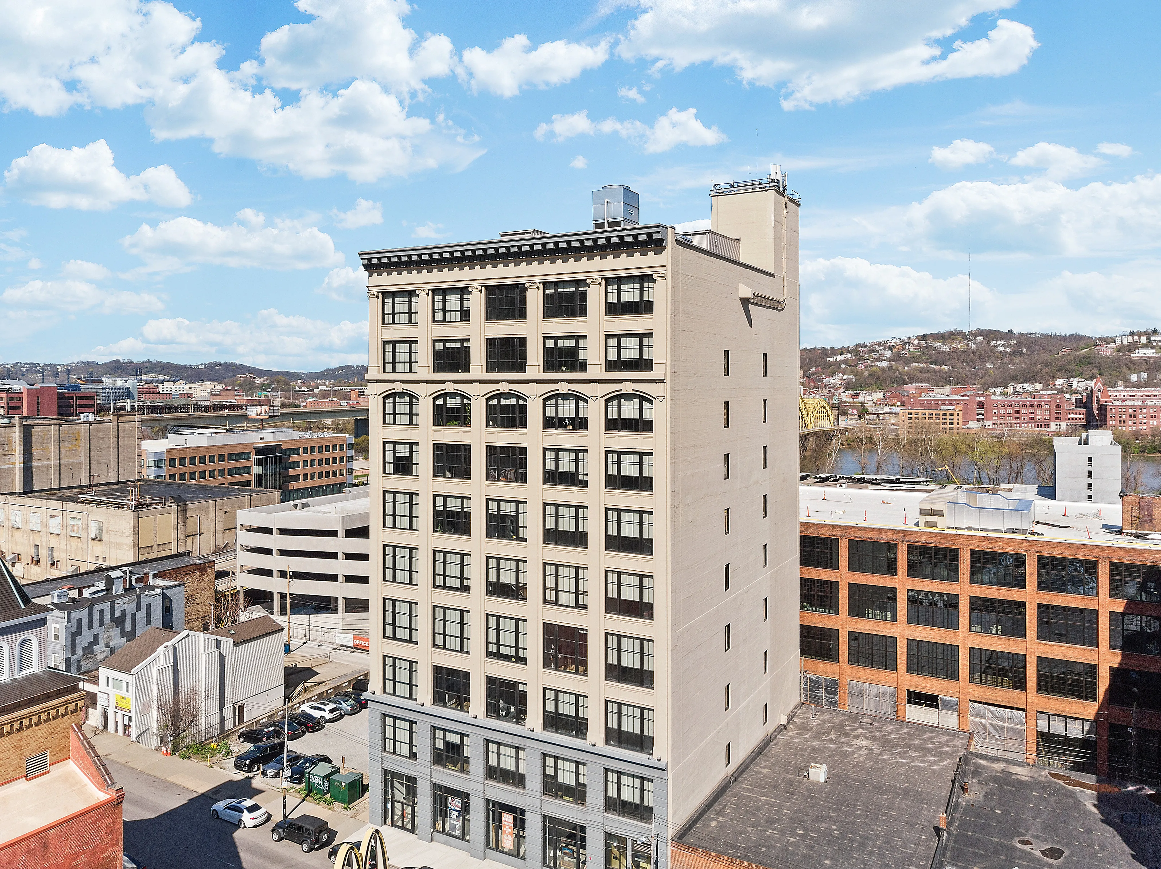 Modern high-rise building with large windows and stunning river views, set against a vibrant urban backdrop with hills and blue skies.