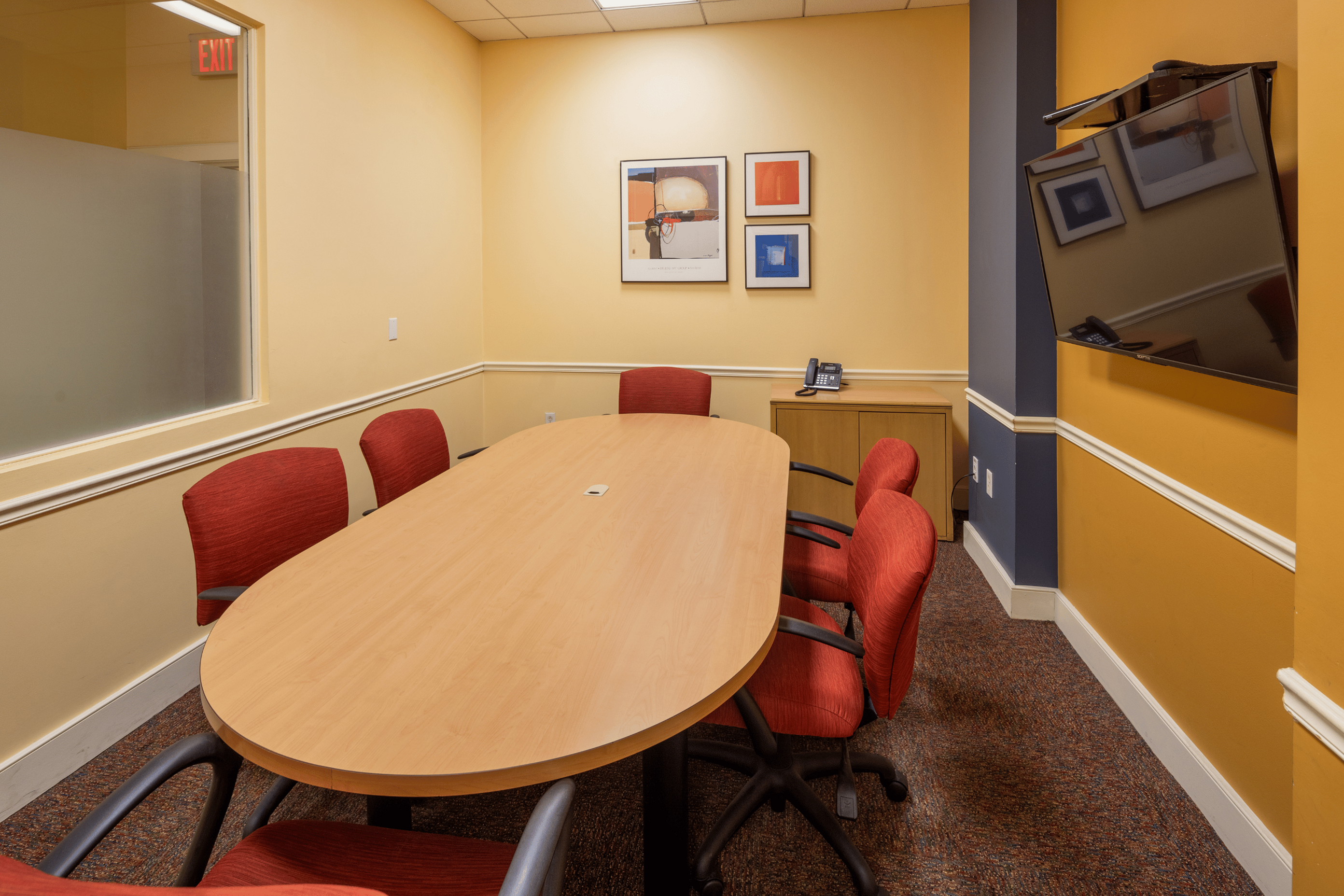 Contemporary meeting room with vibrant red chairs, a sleek wooden table, and modern artwork on sunny yellow walls.