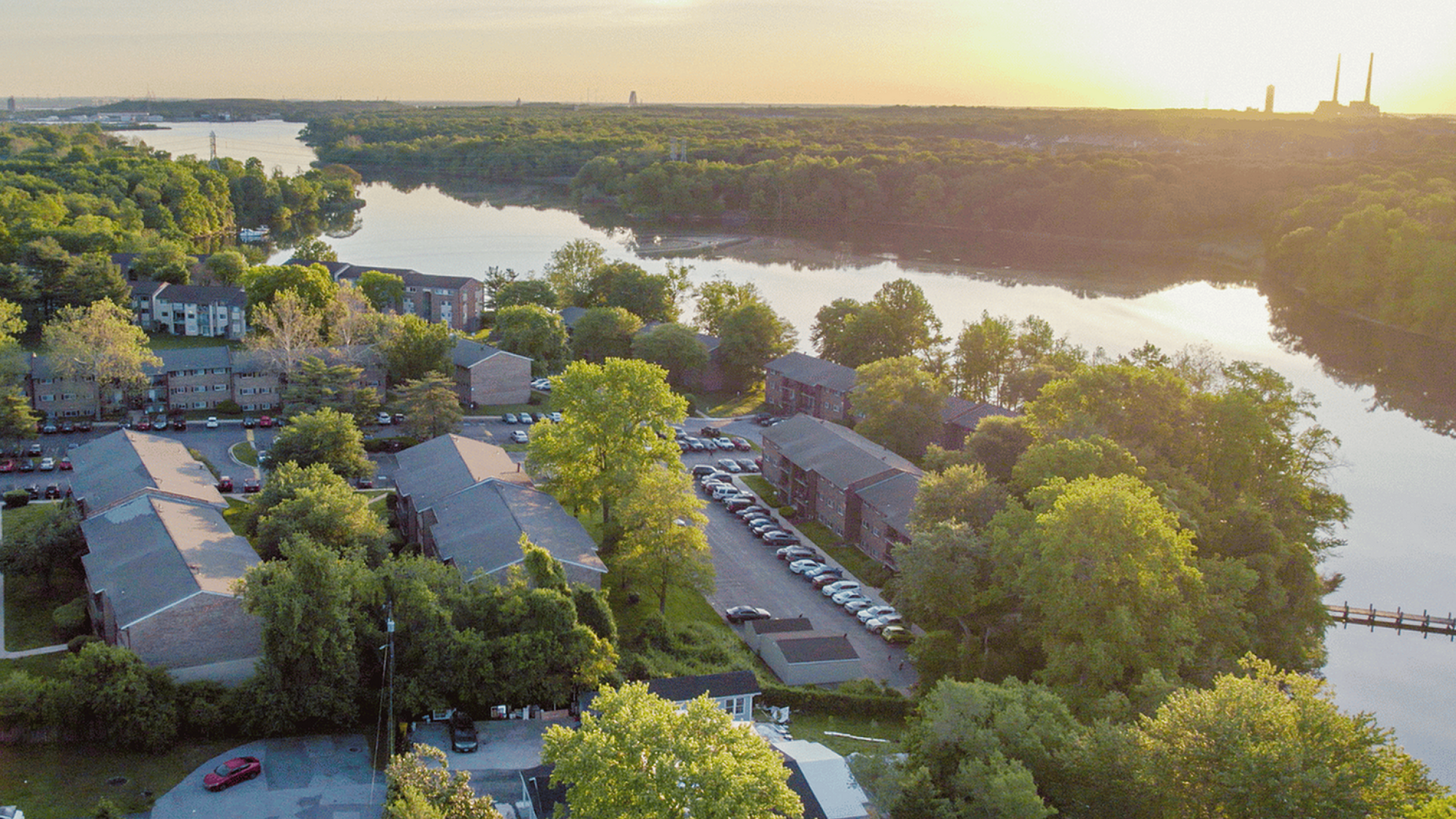 Aerial view of a charming riverside apartment community surrounded by lush greenery during sunset.