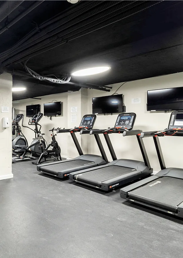 Modern gym facility with treadmills, exercise bikes, weight machines, and wall-mounted TVs.