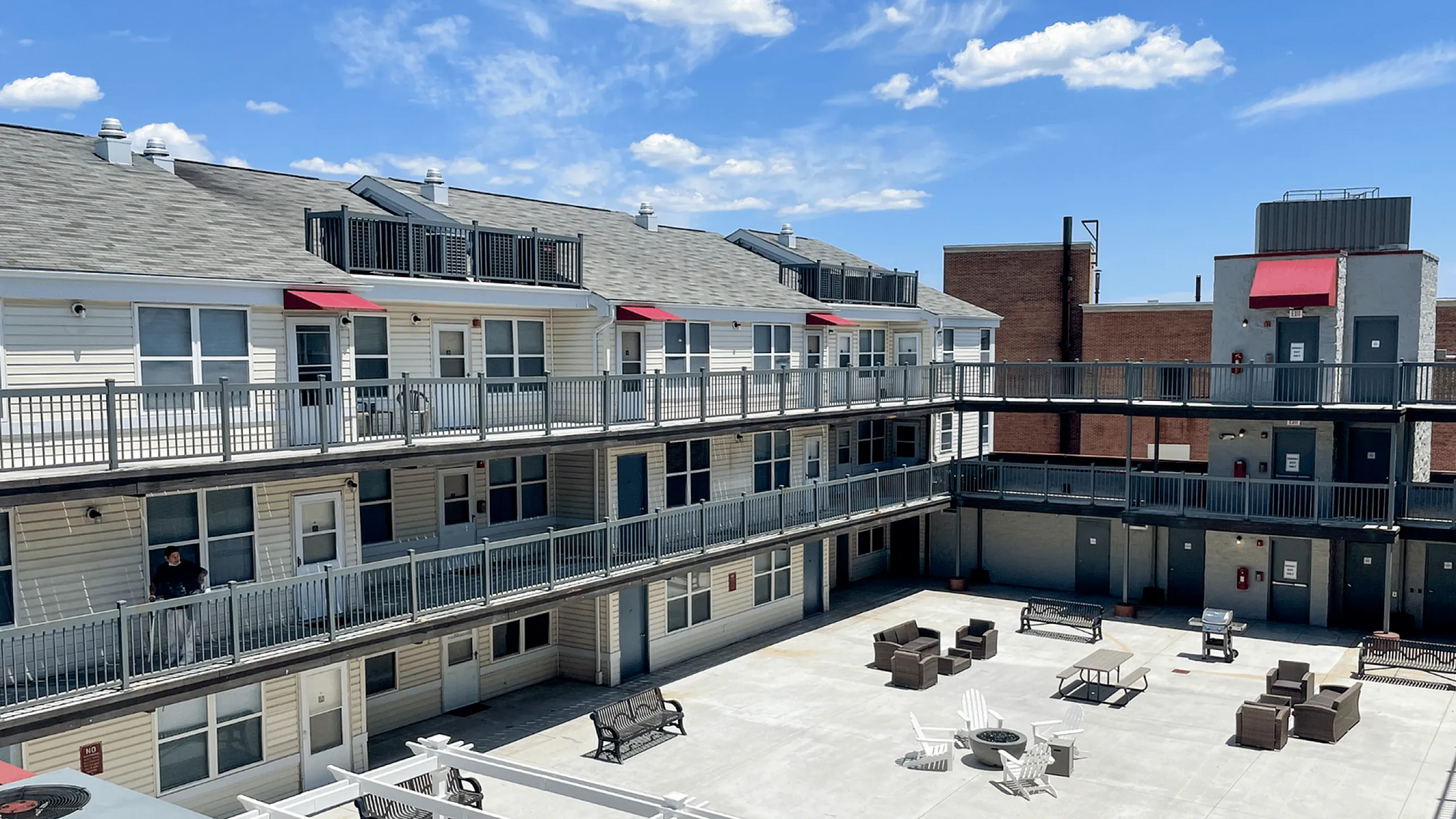 Apartment complex with open courtyard featuring seating areas and modern railings under a bright blue sky.
