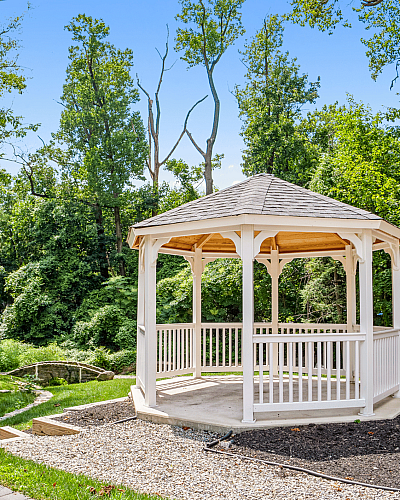 Picturesque white wooden gazebo in a lush green garden, surrounded by tall trees and a serene walking path.