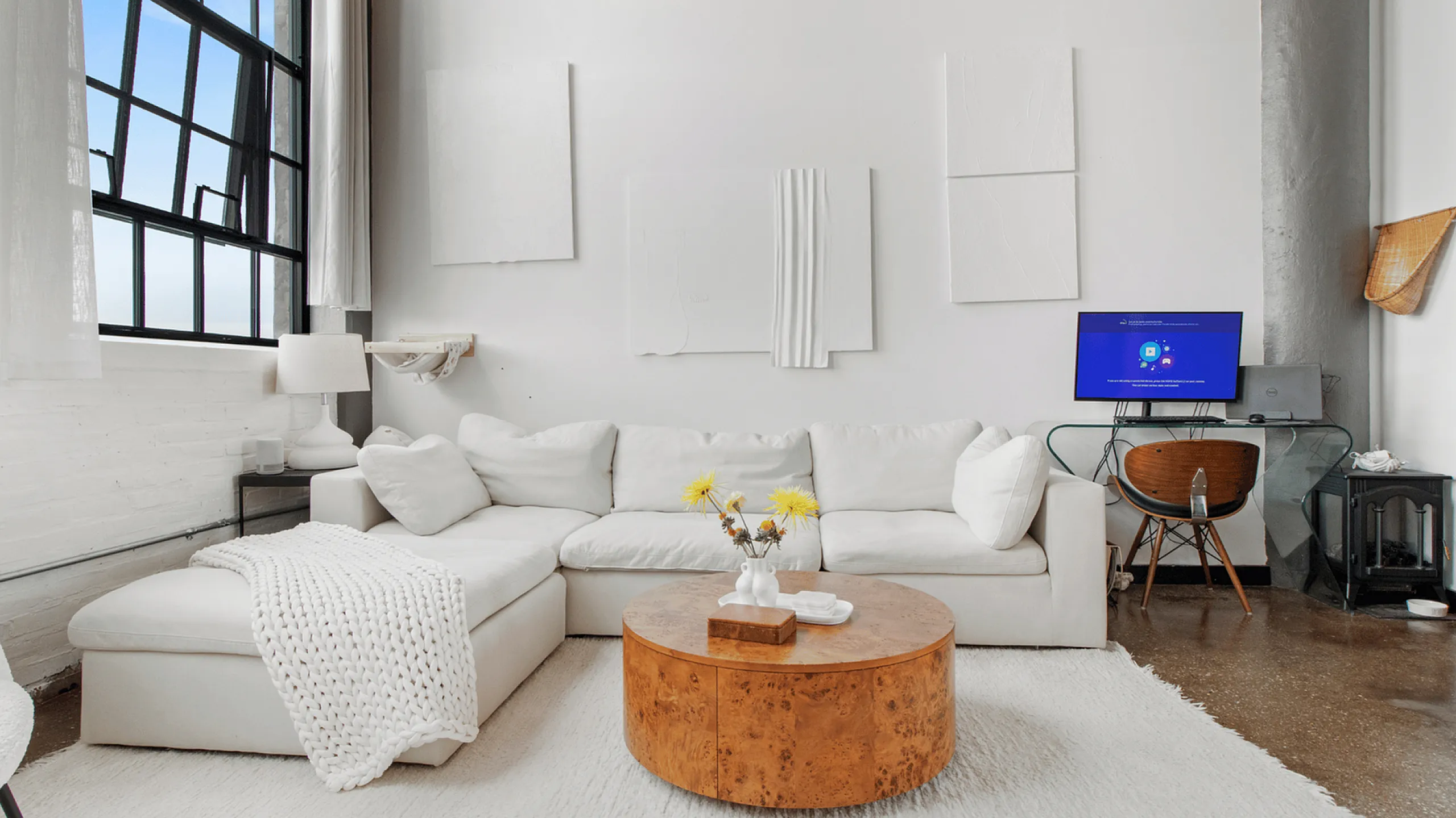 Stylish modern living room with large windows, a cozy white sectional sofa, a round wooden coffee table, and an elegant workspace.