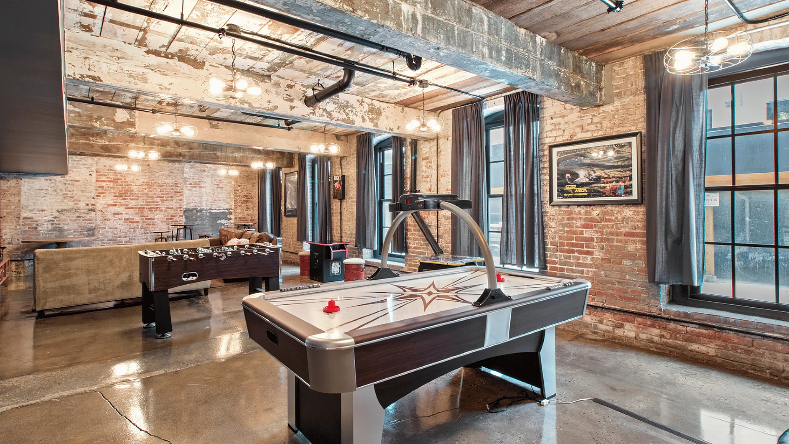 Stylish industrial loft featuring an air hockey and foosball table, with exposed brick walls and large windows.