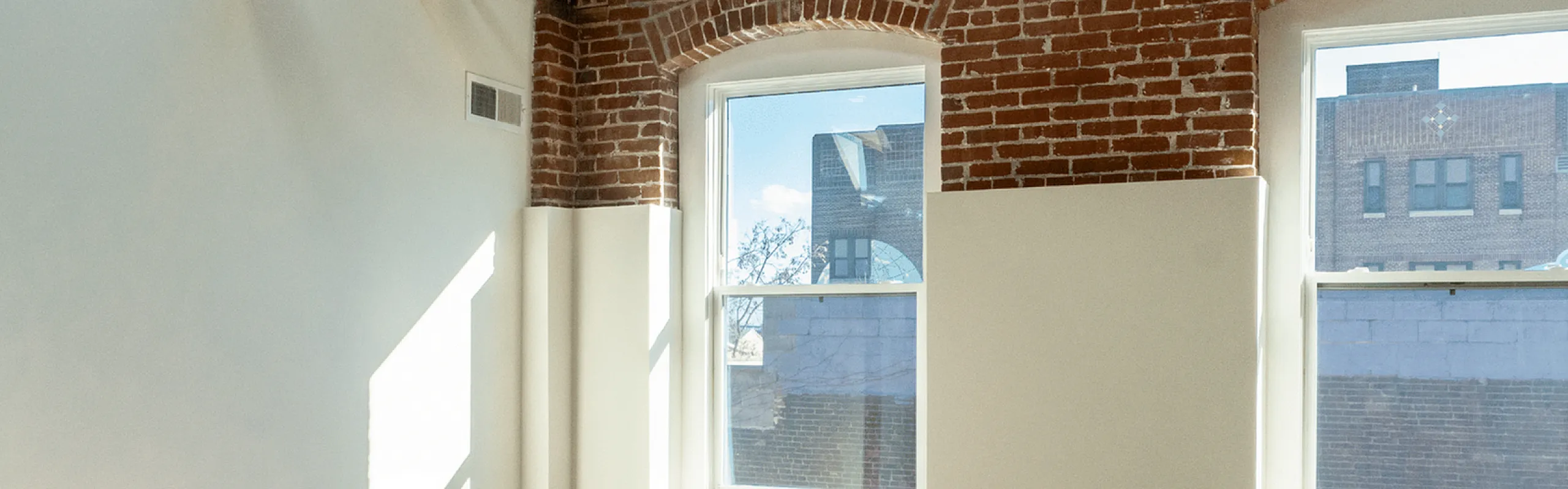 Sunlit loft room with exposed brick walls, wooden beams, and large windows.