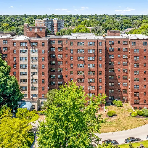 Charming brick apartment building surrounded by lush green trees and vibrant landscaping, offering a serene living environment.