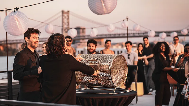 Chic rooftop gathering at sunset with smiling guests and elegant lanterns, overlooking a stunning bridge.