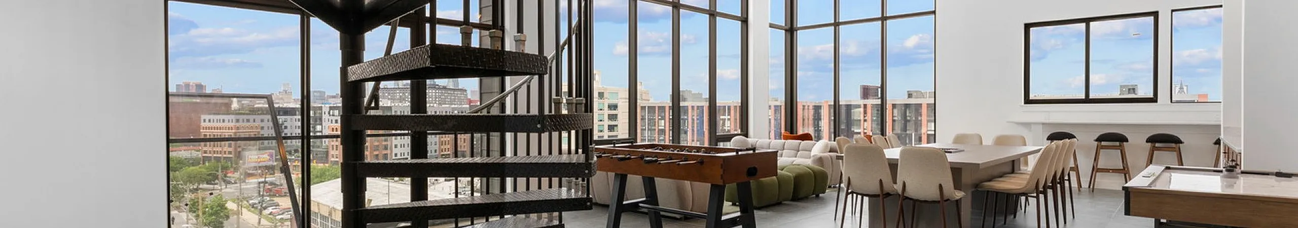 Modern loft space with stunning spiral staircase, large windows offering panoramic city views, chic dining area, and recreational foosball table.