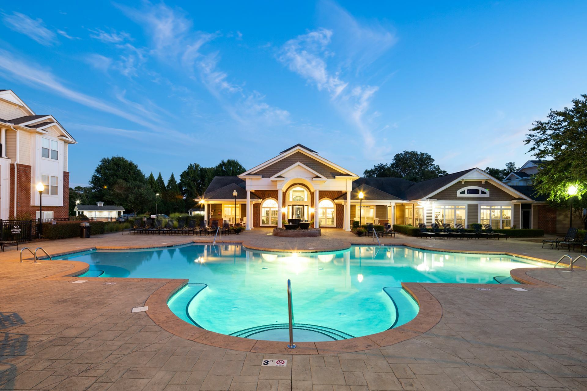 Illuminated swimming pool with a luxurious patio area during dusk, featuring lounge seating and a grand clubhouse with bright arched windows and columns at Abberly Woods Apartments in Charlotte, NC