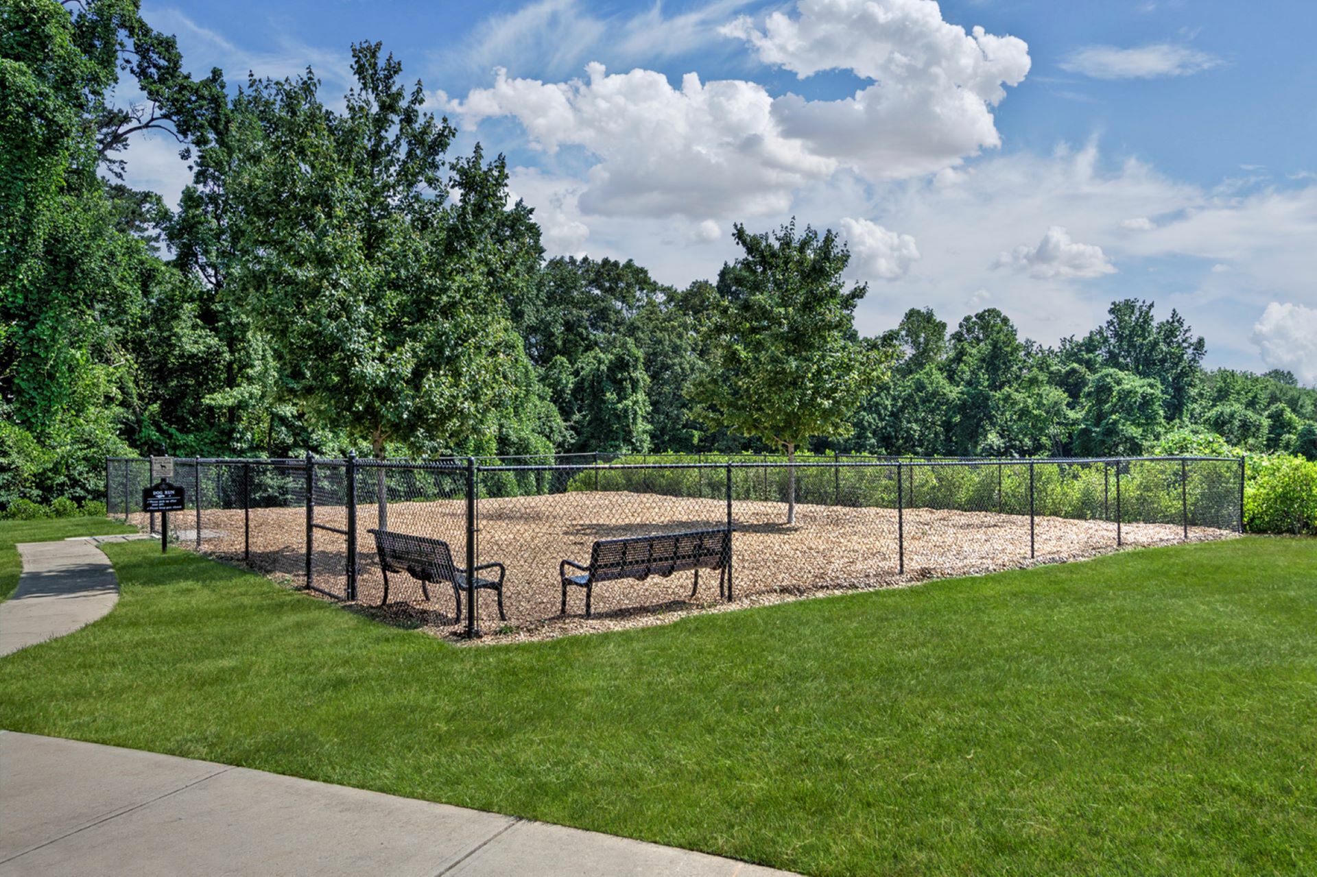 Fenced on-site dog park featuring a spacious play area with mulch ground cover, two benches for pet owners, and surrounded by lush greenery at Abberly Woods Apartment Homes in North Carolina
