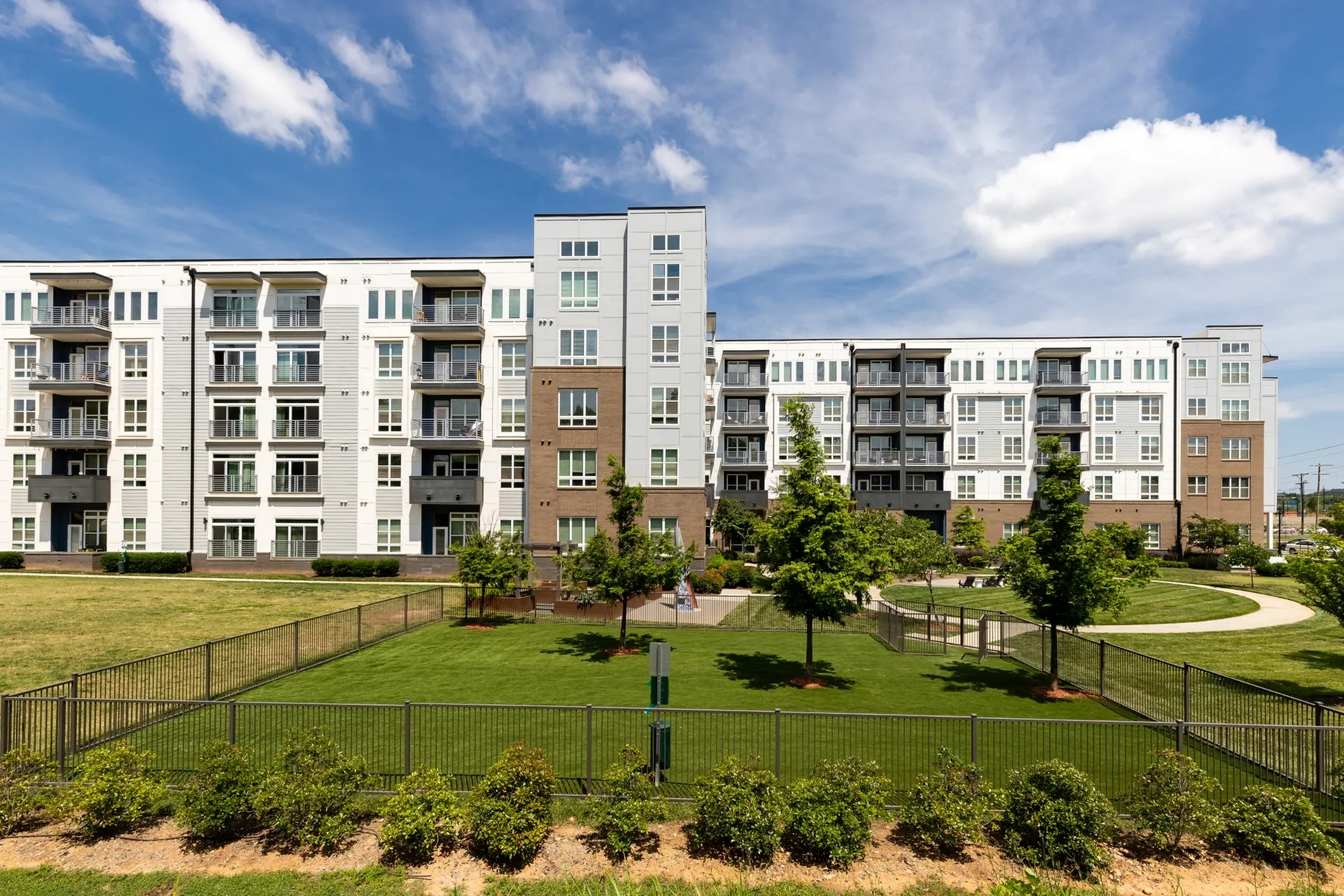 Modern apartment complex with spacious balconies, surrounded by lush greenery and open grass areas, under a clear blue sky.