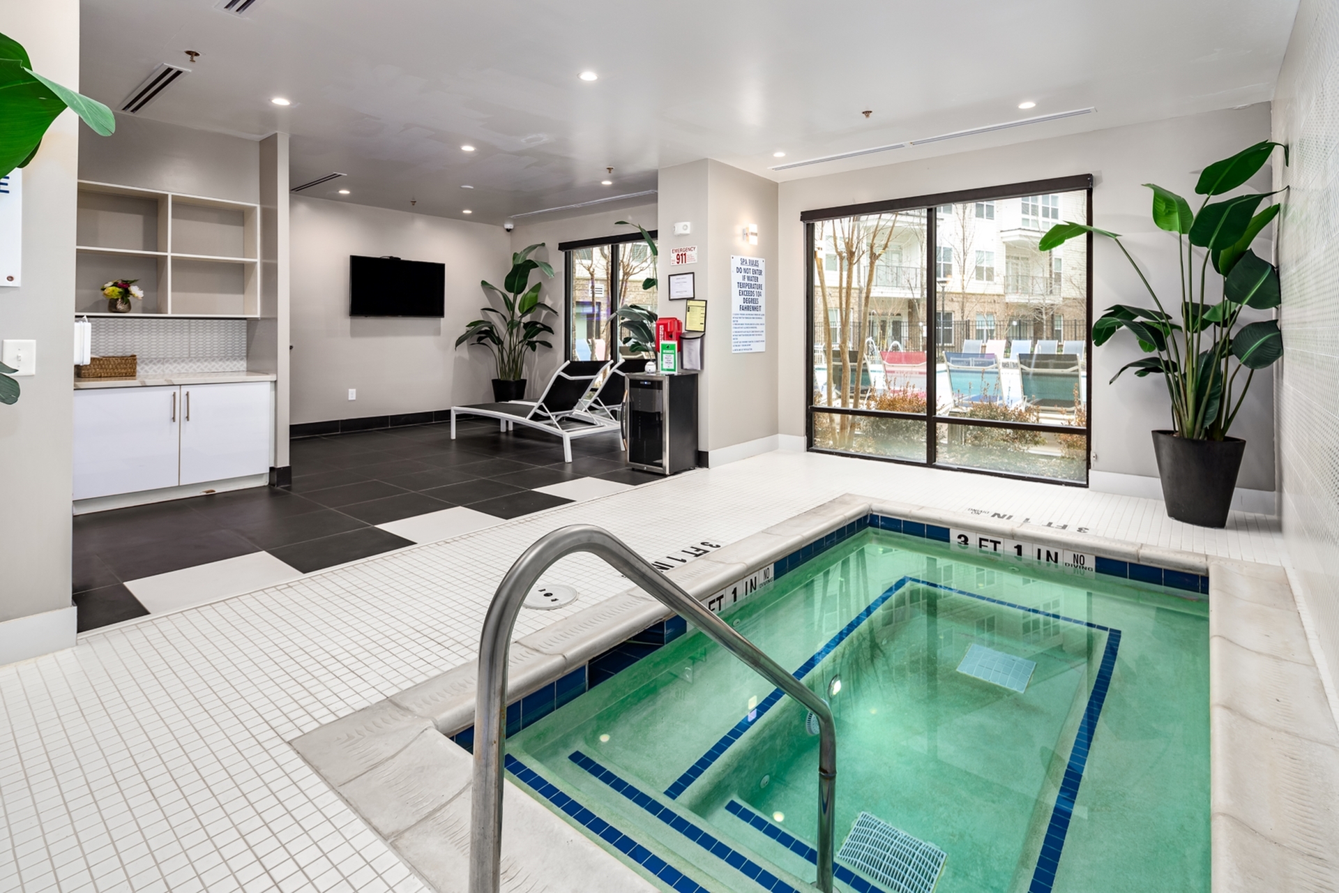 Modern indoor apartment spa area with a luxurious hot tub, sleek lounge chairs, and lush greenery. Large windows offer an inviting view to the outside.
