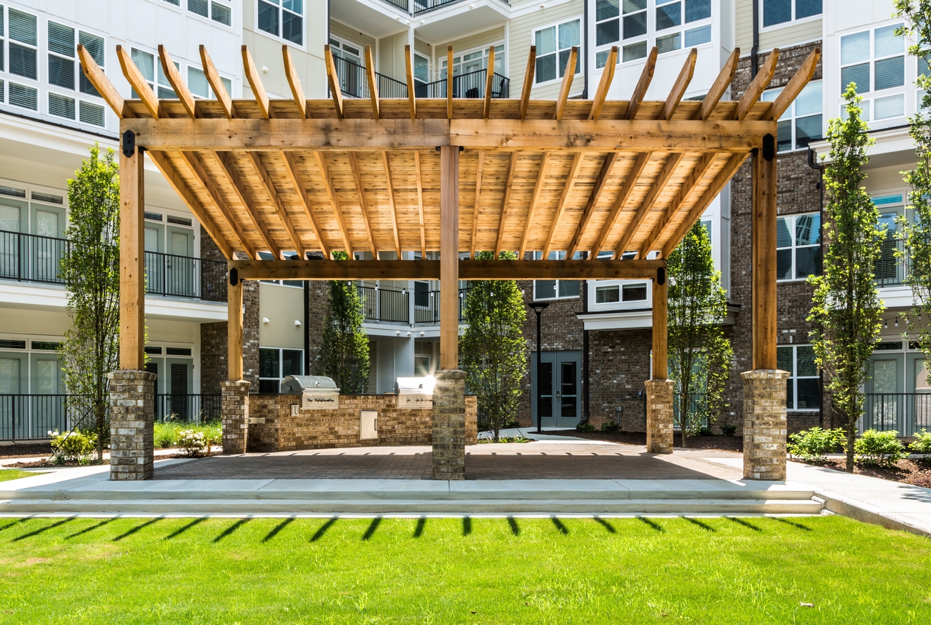 Elegant outdoor communal area featuring a stylish wooden pergola with built-in grills, surrounded by lush greenery in a modern apartment complex.