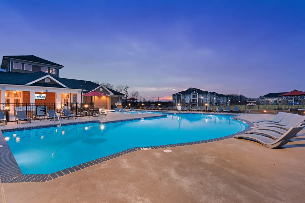 Luxurious apartment complex featuring a sparkling pool with lounge chairs, surrounded by modern buildings at twilight.