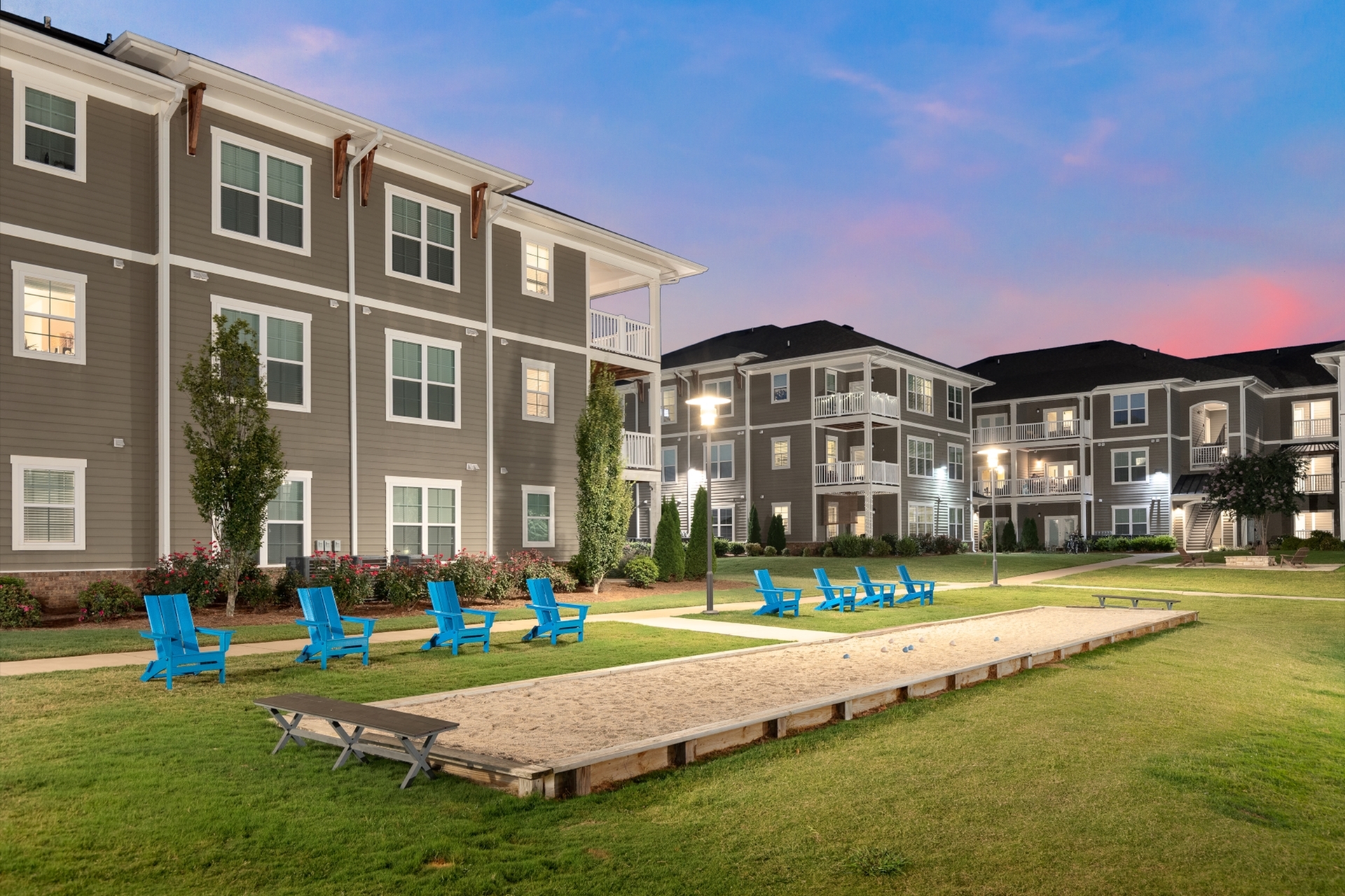 Charming apartment complex at dusk with modern architecture, surrounded by lush greenery and vibrant blue seating, perfect for relaxation and community gatherings.